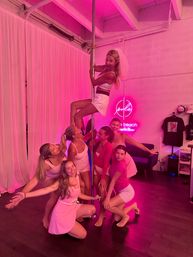 Bachelorette party at a pole-fitness studio: group of women in pink athletic outfits posing playfully around a chrome pole under pink neon lighting