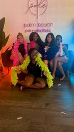 Five women wearing colorful feather boas smiling and posing in a Palm Beach pole and aerial fitness studio in front of a neon wall sign