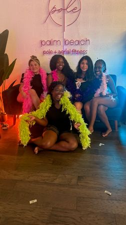 Five women wearing colorful feather boas smiling and posing in a Palm Beach pole and aerial fitness studio in front of a neon wall sign
