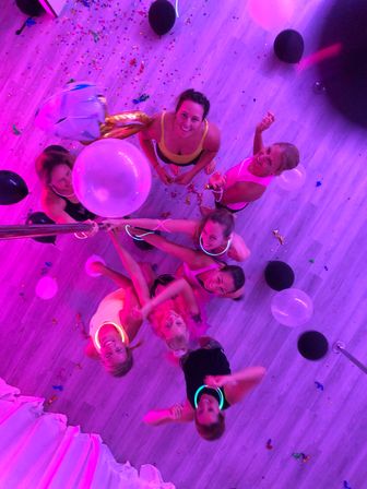 Aerial view of a group of women in a pink-lit indoor dance studio celebrating with balloons, confetti, neon glow necklaces and a dance pole