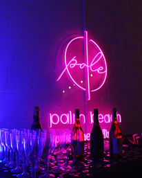 Vibrant pink and purple neon sign reading 'pole' glowing over a party table with rows of champagne flutes, wine bottles, confetti and moody club-style lighting.