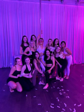 Group of women in activewear posing around a pole in a purple-lit fitness studio, holding play money and party props for a girls' night photo