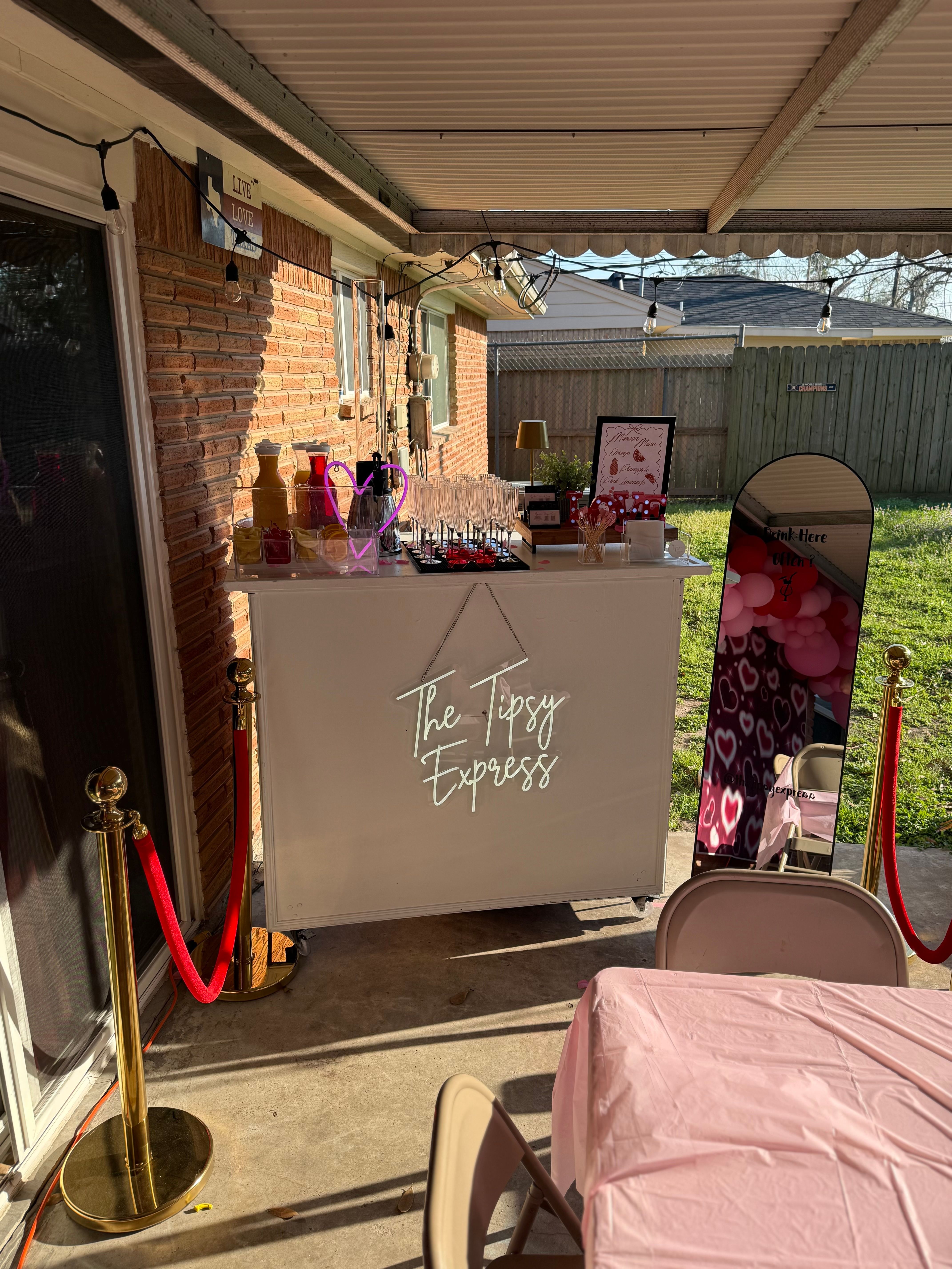 Covered backyard patio party scene with a white mobile bar, champagne flutes, mirror photo booth with pink heart balloons, string lights and red velvet ropes.