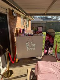 Covered backyard patio party scene with a white mobile bar, champagne flutes, mirror photo booth with pink heart balloons, string lights and red velvet ropes.