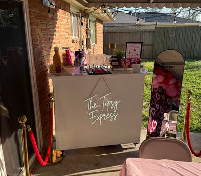 Covered backyard patio party scene with a white mobile bar, champagne flutes, mirror photo booth with pink heart balloons, string lights and red velvet ropes.