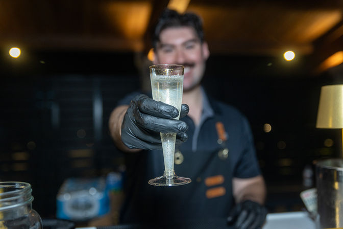 Close-up of a gloved bartender offering a bubbly champagne flute over a bar counter in a dimly lit nightlife setting — cheers!