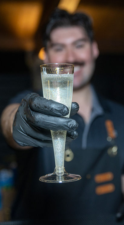 Close-up of a gloved bartender offering a bubbly champagne flute over a bar counter in a dimly lit nightlife setting โ cheers!
