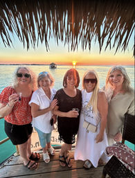 Five smiling women on a tropical wooden dock under a palapa, holding drinks at sunset over calm turquoise water with a distant thatched boat.
