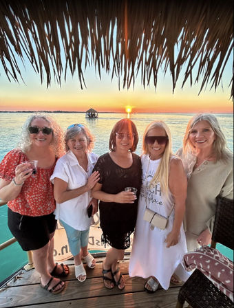 Five smiling women on a tropical wooden dock under a palapa, holding drinks at sunset over calm turquoise water with a distant thatched boat.