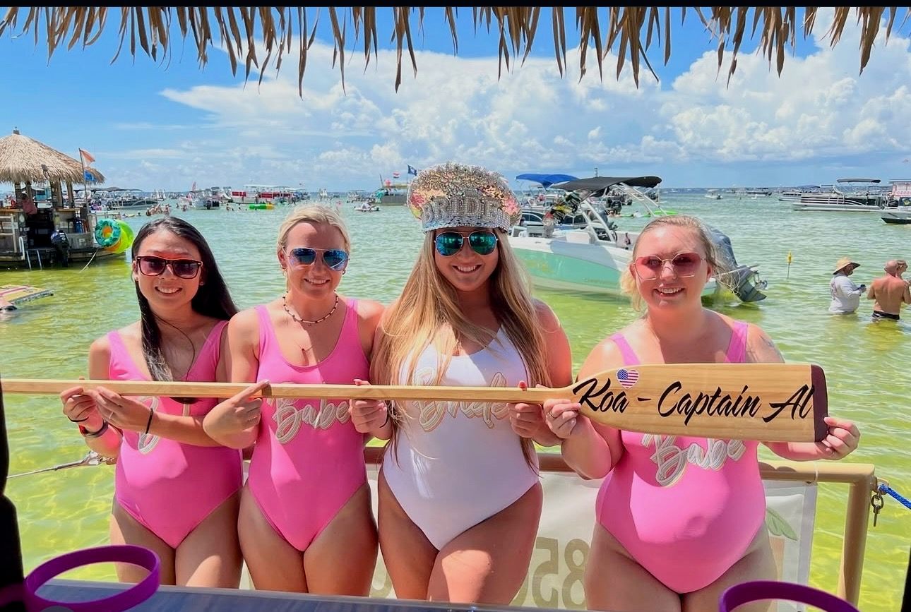 Four women in matching pink and white swimsuits smile under a thatched tiki shade on a sunny beach, holding a wooden paddle with writing, with anchored boats and shallow turquoise water behind them.
