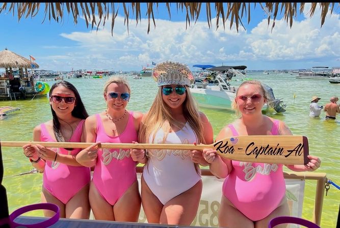 Four women in matching pink and white swimsuits smile under a thatched tiki shade on a sunny beach, holding a wooden paddle with writing, with anchored boats and shallow turquoise water behind them.