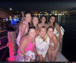 Seven friends in swimsuits smiling and laughing on a waterfront dock at night, tiki-bar lights and marina buildings reflecting on calm water