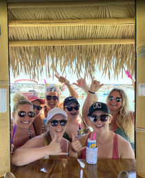 Group of smiling women in sunglasses giving shaka signs and holding canned drinks at a thatched tiki bar overlooking a crowded sunny beach with boats — summer beach party vibe.
