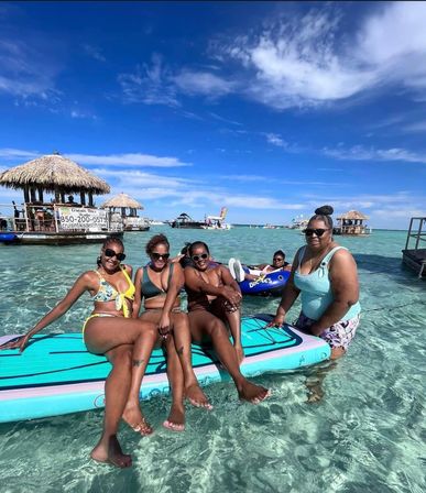 Four friends in swimsuits relaxing on a turquoise paddleboard in crystal-clear shallow tropical water with thatched-roof tiki huts, boats and a bright blue sky in the background.