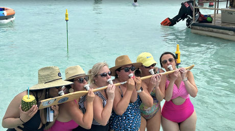 Seven women in swimsuits and sun hats stand in shallow turquoise water sharing a wooden shot-ski and sipping shots, one holding a pineapple cup near a dock