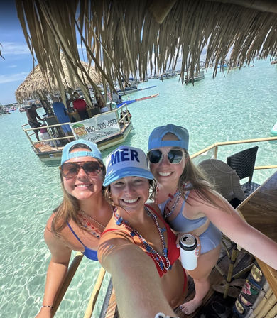Three smiling women in sun hats and bikinis on a floating tiki bar over crystal-clear turquoise shallow water with boats and thatched-roof huts in the background