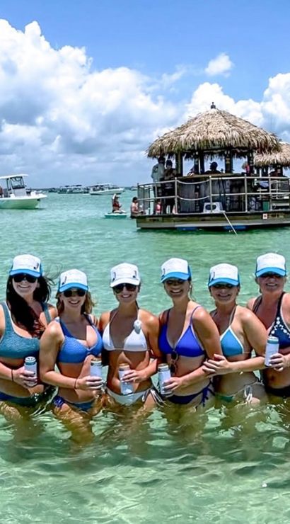 Six women in matching blue-and-white caps and bikinis standing waist-deep on a shallow turquoise sandbar, holding canned drinks with tiki-style floating bars and boats in the background under a sunny, partly cloudy sky.