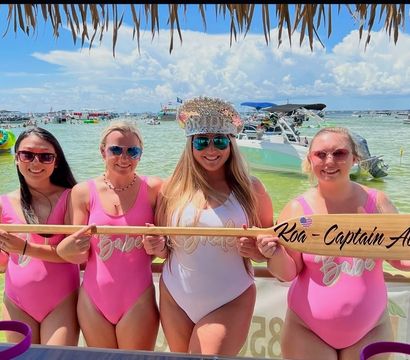 Four women in matching pink and white swimsuits smile under a thatched tiki shade on a sunny beach, holding a wooden paddle with writing, with anchored boats and shallow turquoise water behind them.