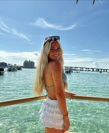 Smiling young woman in a striped bikini top and white ruffled skirt leans on a railing over clear turquoise water with boats, a bridge and sunny blue sky behind her.