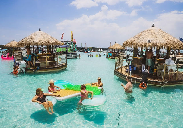 Sunny tropical lagoon with people on inflatable loungers and in shallow turquoise water around thatched-roof floating tiki bars and boats, lively beach-party vibe.