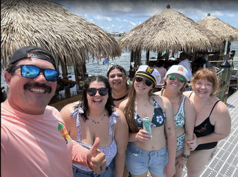 Fun group selfie of six smiling adults in swimsuits on a sunny waterfront dock with thatched tiki-hut bars, calm bay water and canned drinks — tropical summer boater vibe.