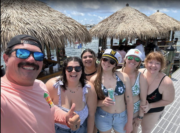 Fun group selfie of six smiling adults in swimsuits on a sunny waterfront dock with thatched tiki-hut bars, calm bay water and canned drinks — tropical summer boater vibe.