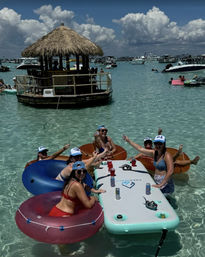 Group on colorful inner tubes around an inflatable floating table in clear turquoise shallow water at a crowded sandbar with a thatched-roof tiki bar and anchored boats — sunny boat party vibe.