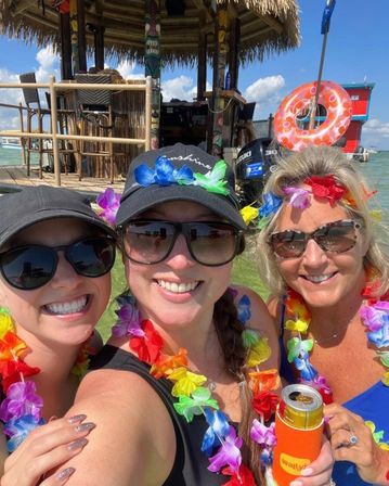 Three smiling women in sunglasses wearing colorful leis take a selfie in shallow turquoise water in front of a thatched tiki hut and floating bar, holding a canned drink on a sunny day.