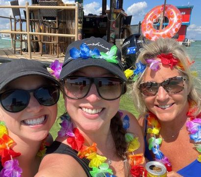 Three smiling women in sunglasses wearing colorful leis take a selfie in shallow turquoise water in front of a thatched tiki hut and floating bar, holding a canned drink on a sunny day.