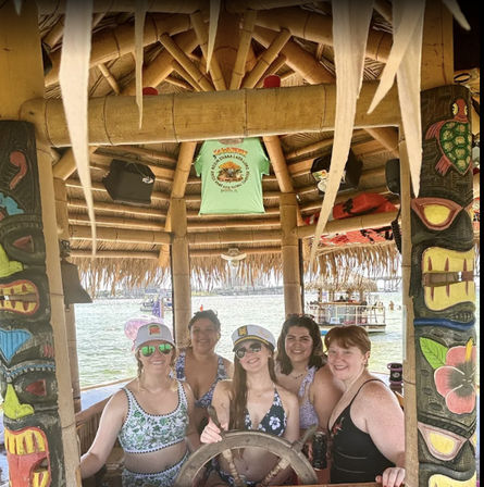 Five women in swimsuits and captain hats posing at the helm of a thatched tiki-style boat bar, framed by colorful carved totem posts with water and a docked boat in the background.