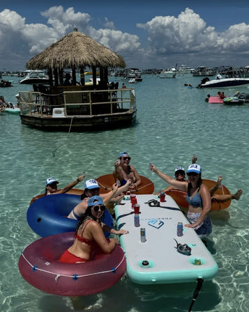 Group on colorful inner tubes around an inflatable floating table in clear turquoise shallow water at a crowded sandbar with a thatched-roof tiki bar and anchored boats — sunny boat party vibe.
