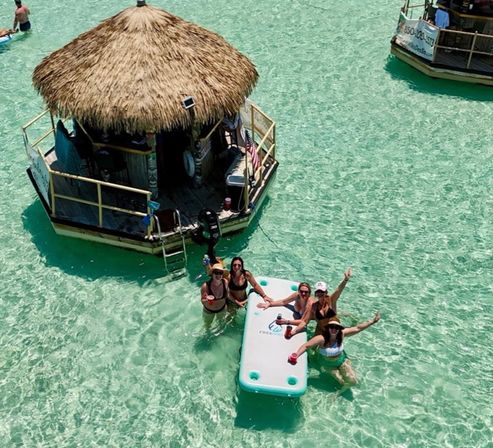 Aerial view of five friends in swimsuits enjoying drinks on a floating aqua platform beside a thatched-roof tiki bar in shallow crystal-clear turquoise tropical water