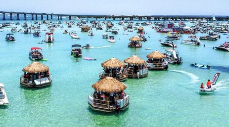 Aerial view of a crowded turquoise coastal bay with dozens of boats and floating tiki-hut party platforms, people enjoying sun and water with a long bridge on the horizon.