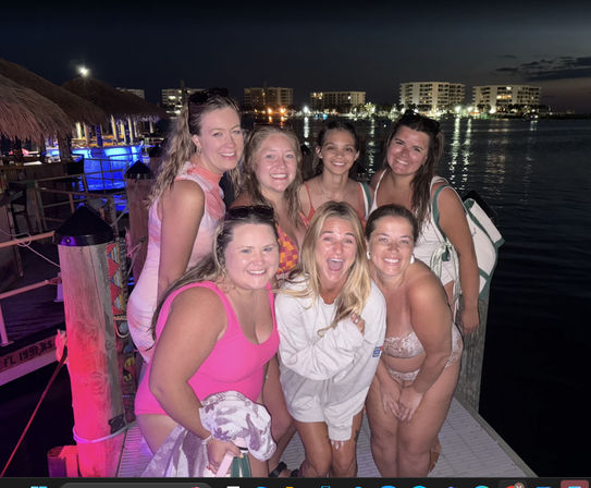 Seven friends in swimsuits smiling and laughing on a waterfront dock at night, tiki-bar lights and marina buildings reflecting on calm water