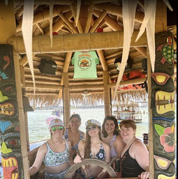 Five women in swimsuits and captain hats posing at the helm of a thatched tiki-style boat bar, framed by colorful carved totem posts with water and a docked boat in the background.