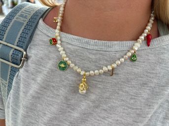 Close-up of a pearl charm necklace with green and red enamel charms, a gold octopus and small initial pendant worn over a light gray T‑shirt beside a blue patterned backpack strap