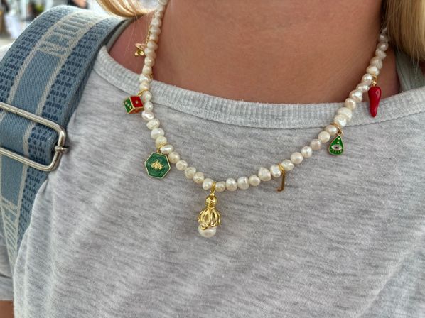 Close-up of a pearl charm necklace with green and red enamel charms, a gold octopus and small initial pendant worn over a light gray T‑shirt beside a blue patterned backpack strap