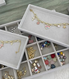 White velvet jewelry display tray with compartments holding a gold charm bracelet with enamel heart and cross charms, starfish and flame motifs, colorful gemstone heart beads, evil-eye beads and assorted studs.