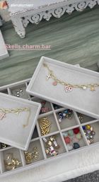 Close-up of a vendor table jewelry display: gray velvet trays holding gold charm bracelets and assorted colorful charms — heart, cross, starfish, evil-eye beads and lightning bolts — on green fabric with a white decorative wooden table edge.