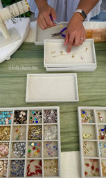 Hands assembling a delicate gold charm bracelet with pliers on a craft-fair table, velvet trays of colorful beads, charms, and pendants arranged nearby.