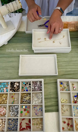Hands assembling a delicate gold charm bracelet with pliers on a craft-fair table, velvet trays of colorful beads, charms, and pendants arranged nearby.