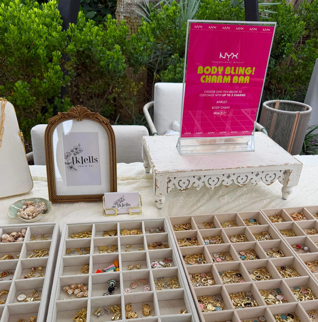 Outdoor charm bar display with a bright pink 'Body Bling! Charm Bar' sign, ornate vintage frame on a white distressed riser, and organized trays filled with gold, enamel and tassel charms for bracelets, anklets and body chains.