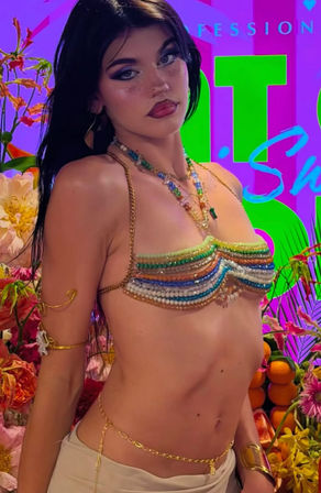 Close-up of a dark-haired model with glitter makeup wearing a rainbow beaded bikini top, layered necklaces and gold body chains against a neon tropical floral backdrop with oranges — festival fashion vibe.