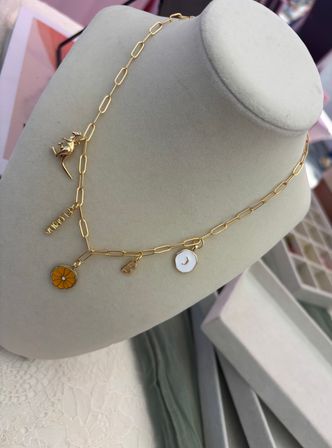 Gold paperclip-link charm necklace on a gray display bust — yellow enamel citrus charm, white moon disc, tiny gold animal charm and small tag, boutique jewelry display.