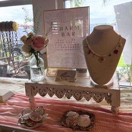 Coastal-themed boutique display with a framed Charm Bar sign, mannequin necklace featuring heart and sun charms, seashell trays, pink roses in a glass vase and hanging chain necklaces on a distressed white pedestal