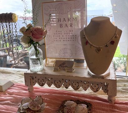 Coastal-themed boutique display with a framed Charm Bar sign, mannequin necklace featuring heart and sun charms, seashell trays, pink roses in a glass vase and hanging chain necklaces on a distressed white pedestal