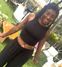 Smiling woman on a grassy outdoor patio wearing a black crop top, shell waist chain, tattoos and sunglasses on her head in a relaxed tropical lounge setting.