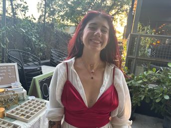 Smiling woman with red hair wearing a red satin top and sheer white blouse, layered necklaces and small chest tattoo, posing by a table of jewelry displays on an outdoor patio bathed in warm sunset light