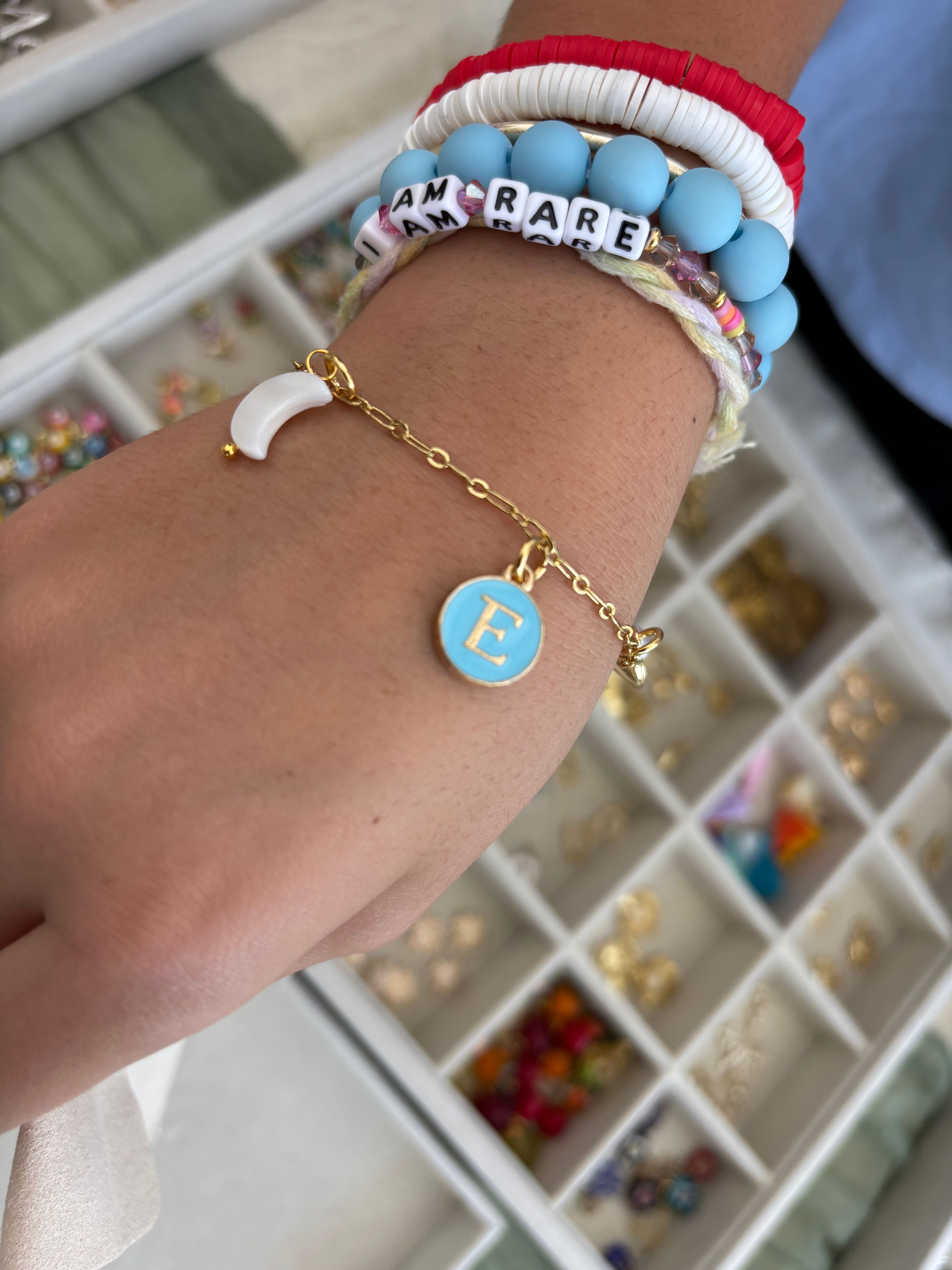 Close-up of wrist wearing stacked colorful beaded bracelets and a gold chain with a blue "E" initial charm, blurred jewelry display tray in background.