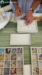 Hands assembling a gold charm bracelet with pliers and tweezers on cream display trays; divided boxes of colorful charms and pendants on a green tablecloth, handmade jewelry display.